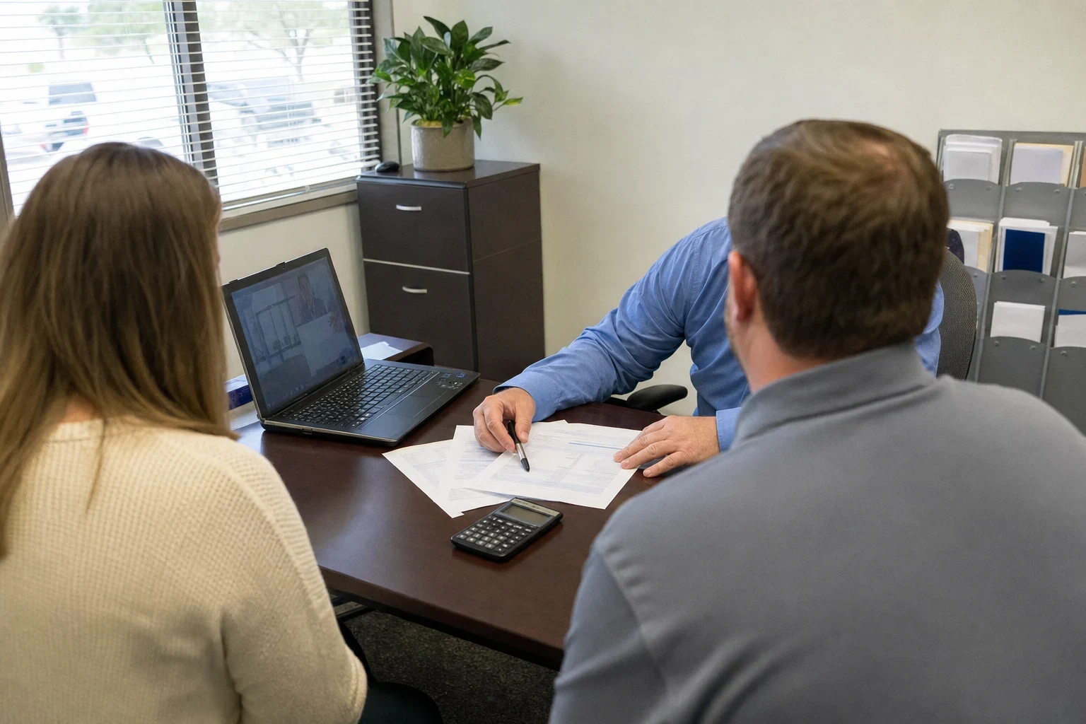 Couple seen from behind sitting at a desk with an agent in a blue shirt who points a pen at printed worksheets beside a calculator and laptop in a small office