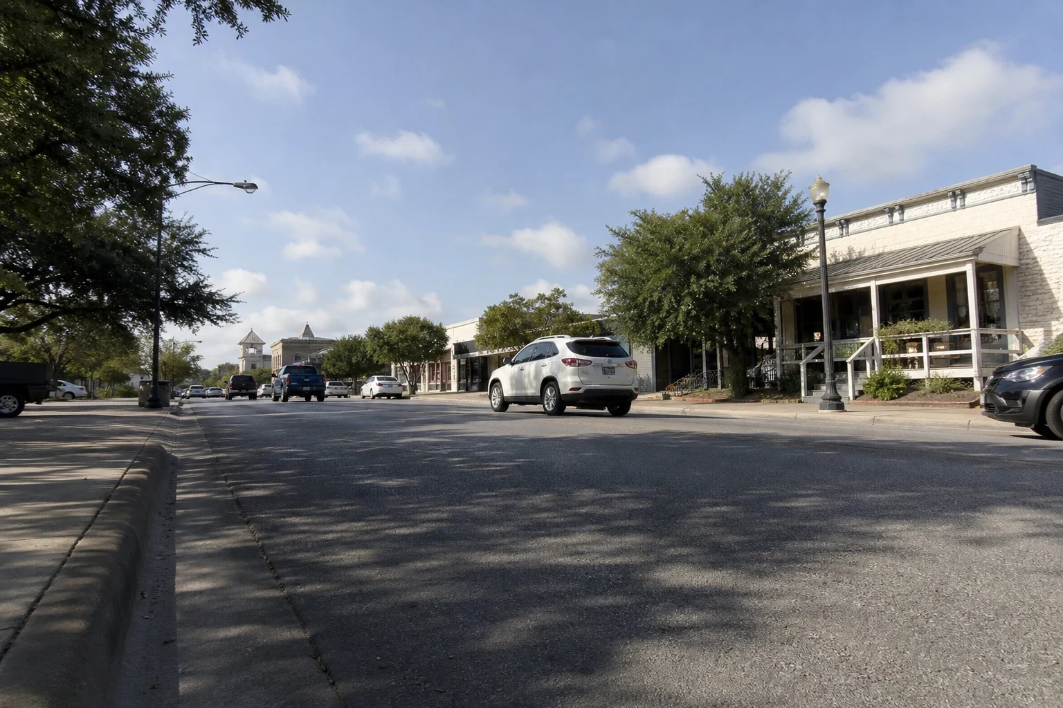 Low-angle view of a small-town Texas main street with a white SUV, parked pickups, shade trees, lamp posts and limestone storefronts under a blue sky