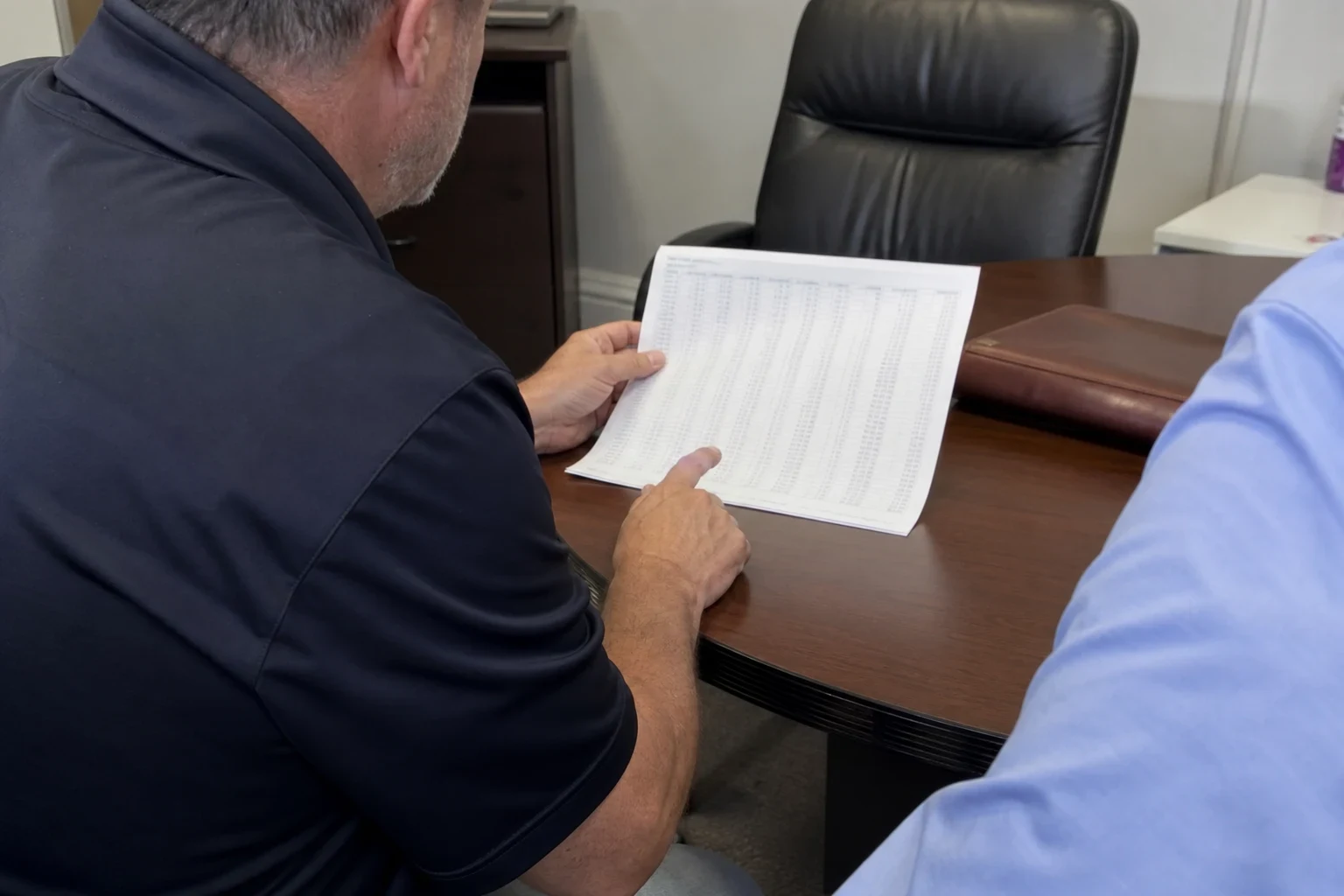 Man in a navy polo points to a printed table of figures at a round wood conference table while a second person in a blue shirt sits beside him in a small office.