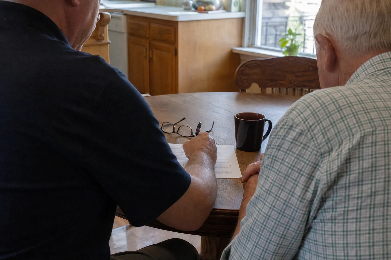 Two men seen from behind at a wooden kitchen table reviewing a paper form, with reading glasses, a pen and a dark coffee mug near oak cabinets and a window