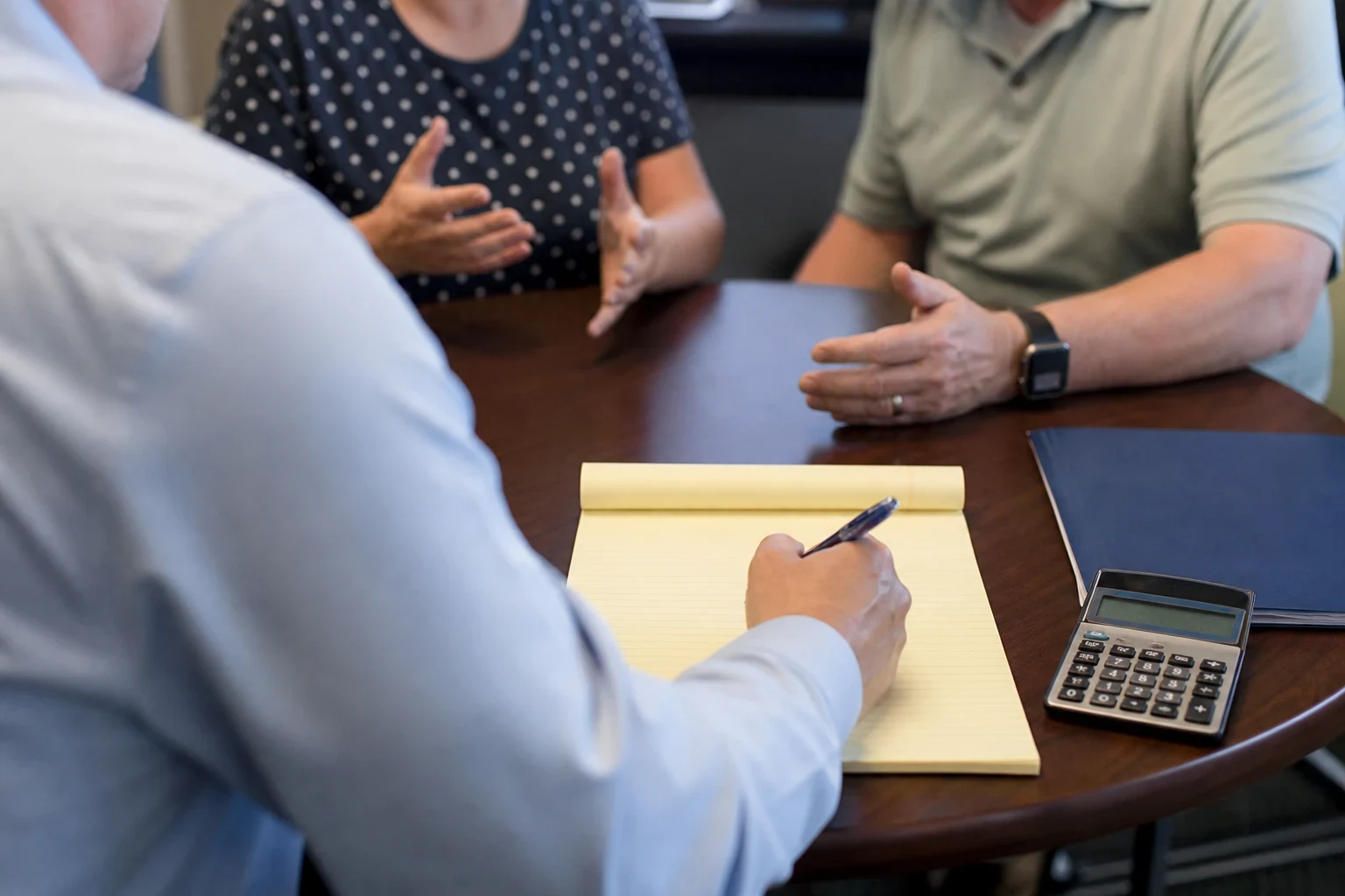 Over-the-shoulder view of an agent writing on a yellow legal pad at a round wooden table as a couple gestures and talks, with a calculator and blue folder nearby